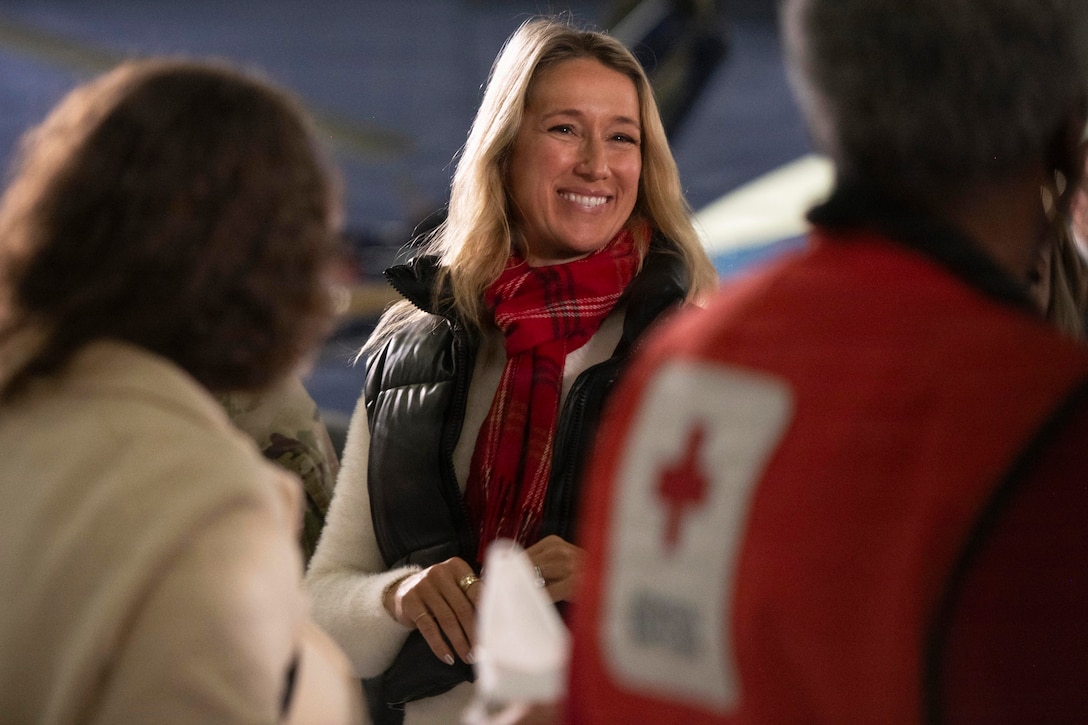 A woman wearing a red scarf, a black winter vest and a white sweater smiles at other people nearby.