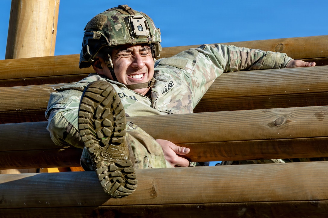 A grimacing soldier wearing a helmet moves through a series of horizontal wooden posts, using a leg and two arms for leverage, on a sunny day outdoors.