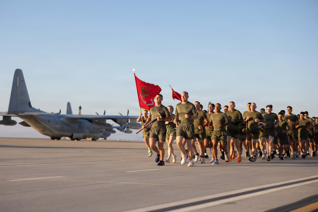 Marines wearing green shorts and T-shirts, one carrying a sledgehammer and two others carrying red flags, run as a group on a flight line during daytime, with a large plane in the background.