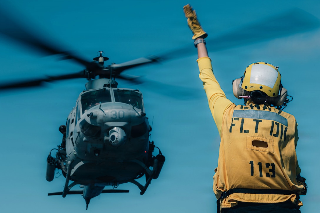 A service member wearing yellow flight-deck attire, a helmet, ear protection and gloves raises one hand to signal a descending helicopter on a sunny day.