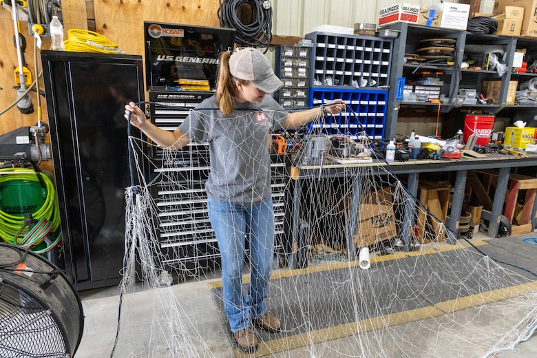A woman in a grey shirt and blue jeans holds up a white fishing net while standing in front of black and blue tool storage carts.