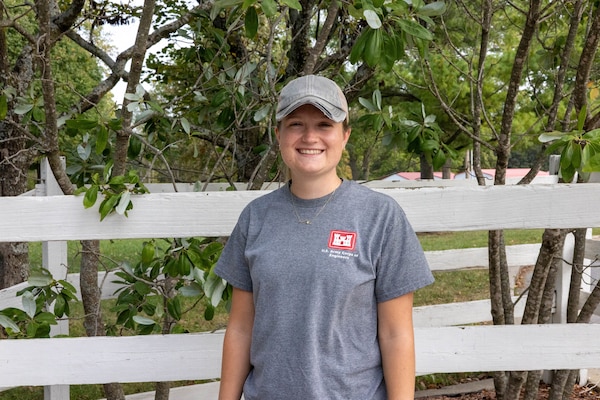 A woman in a grey shirt and baseball hat stands in front of a white fence and green trees and smiles.