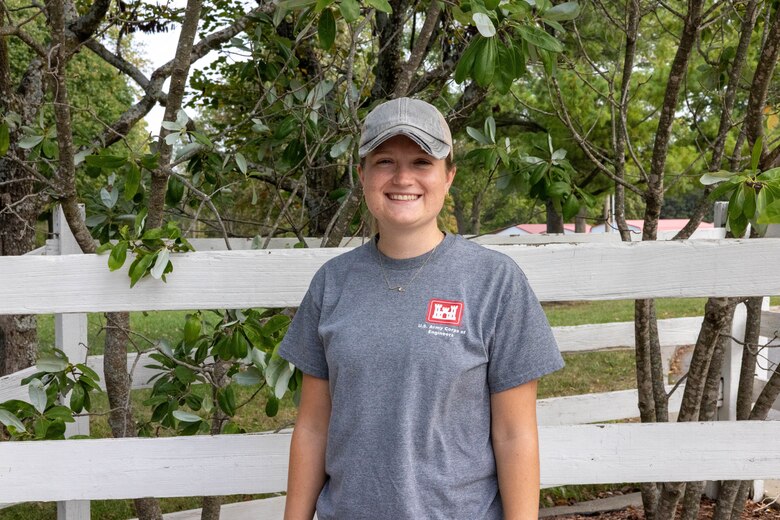 A woman in a grey shirt and baseball hat stands in front of a white fence and green trees and smiles.