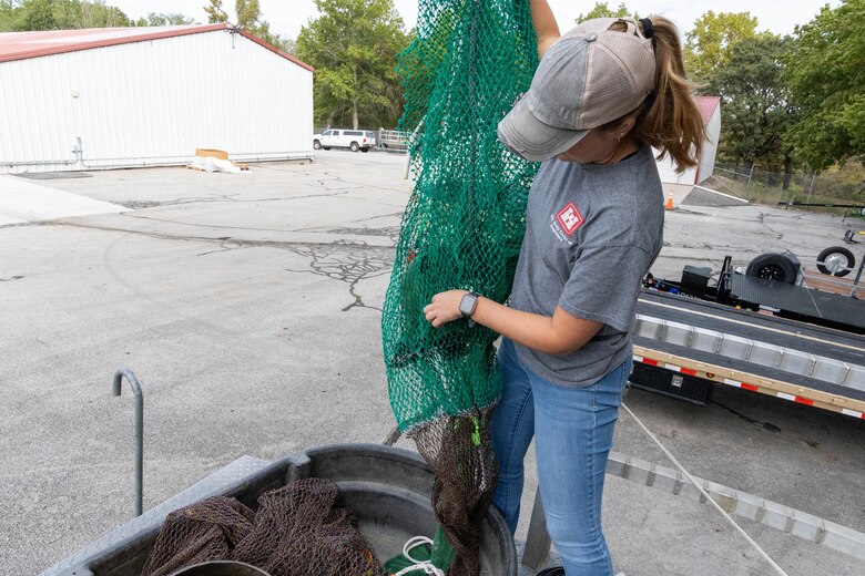 A woman in a grey shirt and blue jeans holds a green net with black asphalt and green trees in the background.