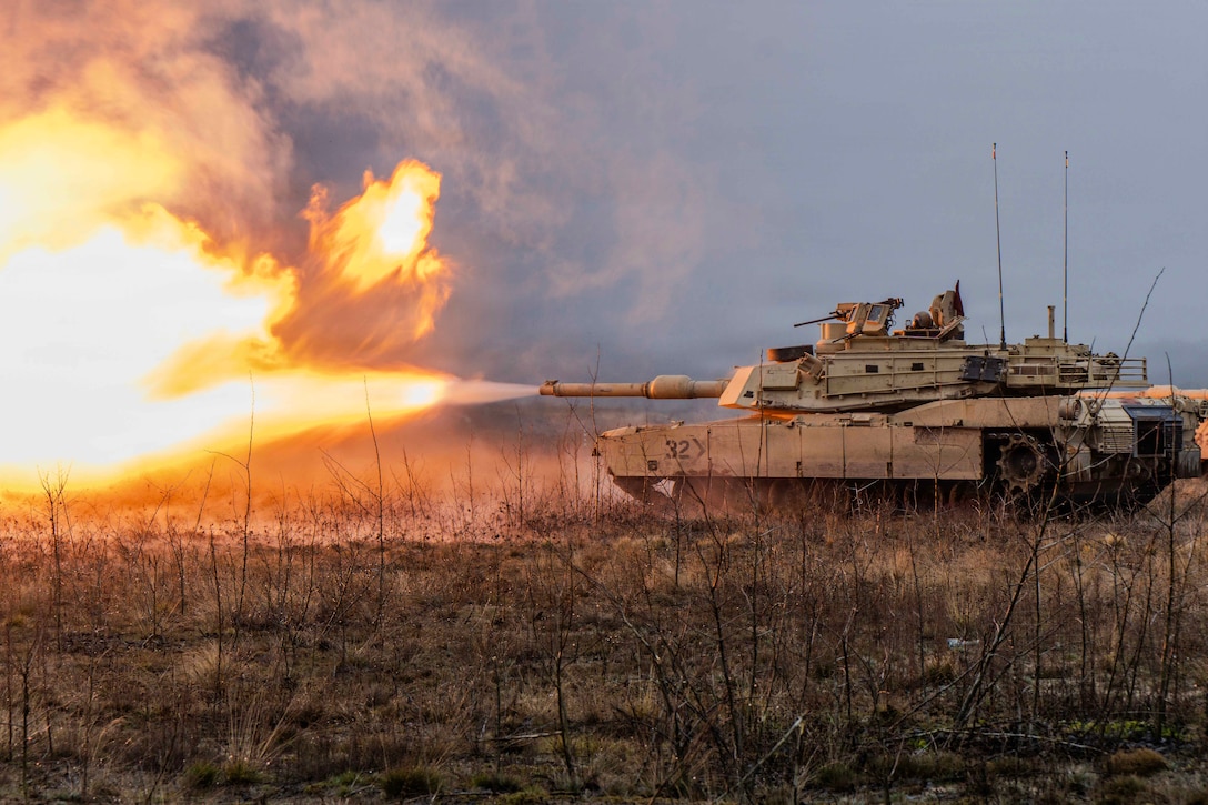 Soldiers operating a tank fire its main gun, creating a burst of fire and swirling smoke in a sparsely vegetated area under a hazy sky.