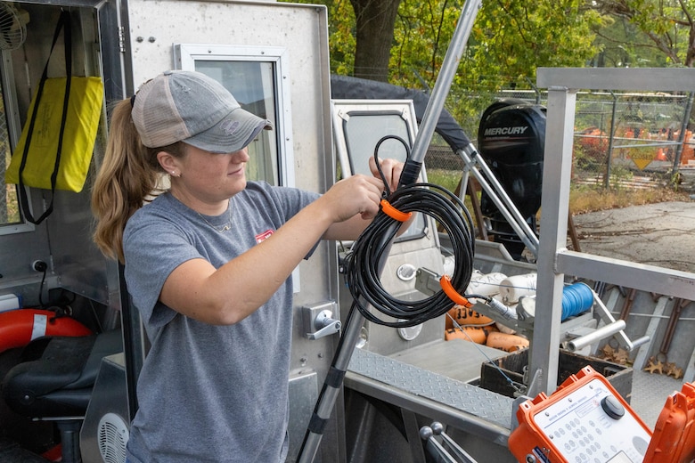 A woman in a grey shirt and baseball hat holds a silver pole with a black cord wound around it.