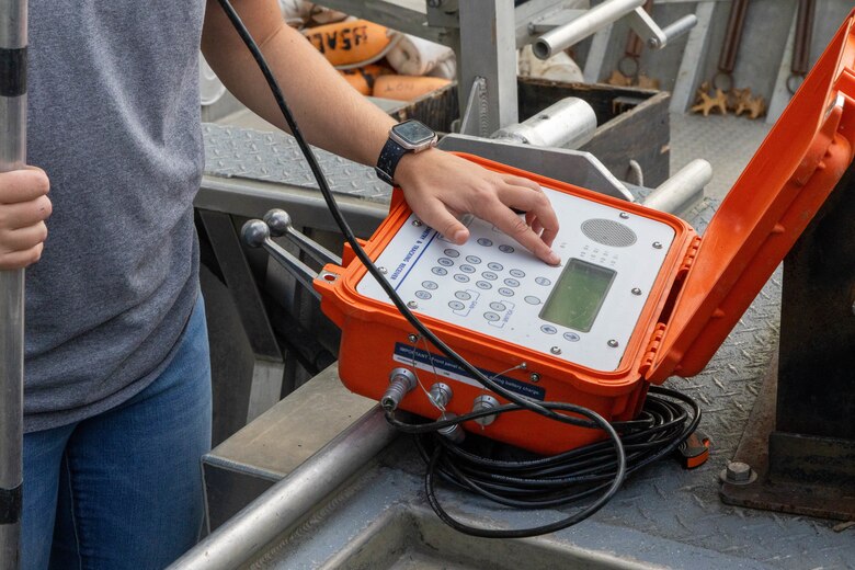 A woman in a grey shirt and blue jeans points to a white screen inside of an orange box while holding a silver pole.
