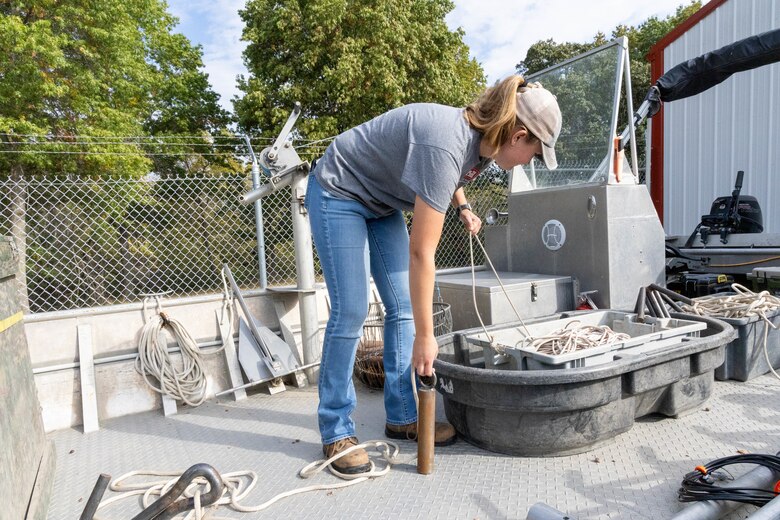 A woman in a grey shirt and blue jeans stands on a grey boat holding a white rope and a brown cylinder with green trees in the background.
