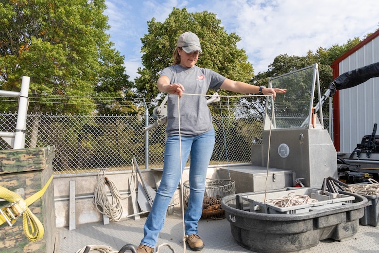 A woman in a grey shirt and blue jeans stands on a boat and holds a white rope with green trees in the background.