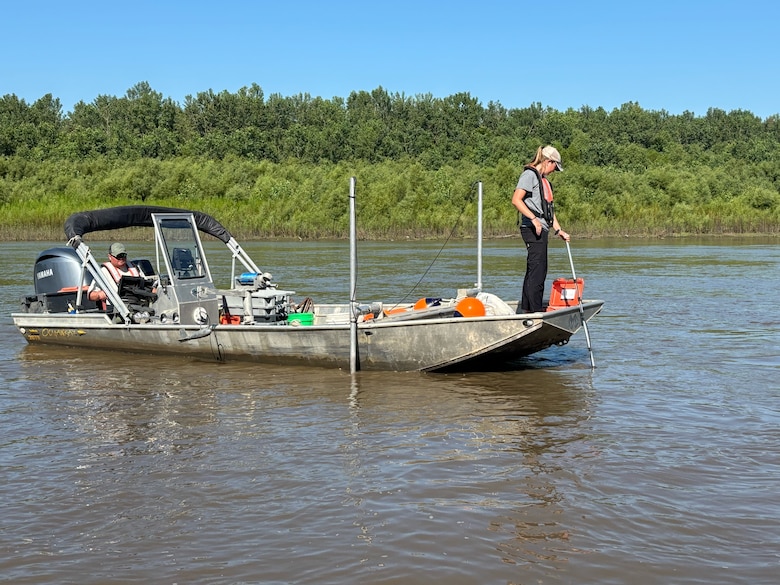 A woman stands on a grey boat holding a white cord with brown water in the forefront and green trees and blue sky in the background.