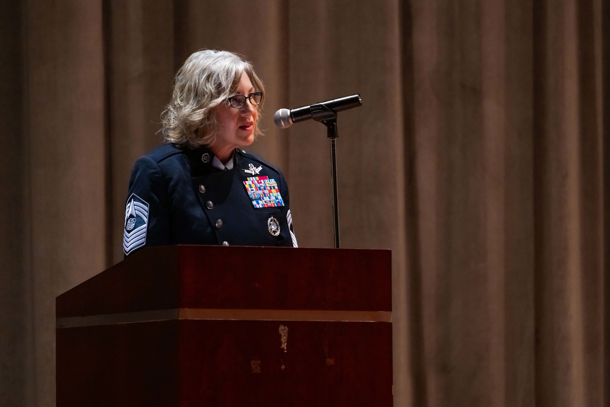 A military chief master sergeant in a blue uniform speak into a microphone at a podium inside an auditorium.