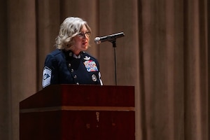 A military chief master sergeant in a blue uniform speak into a microphone at a podium inside an auditorium.