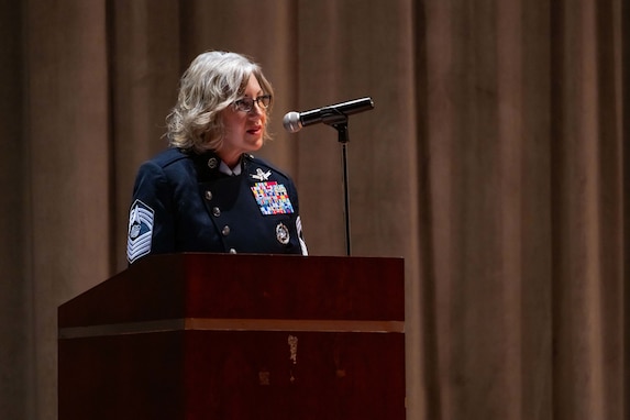A military chief master sergeant in a blue uniform speak into a microphone at a podium inside an auditorium.