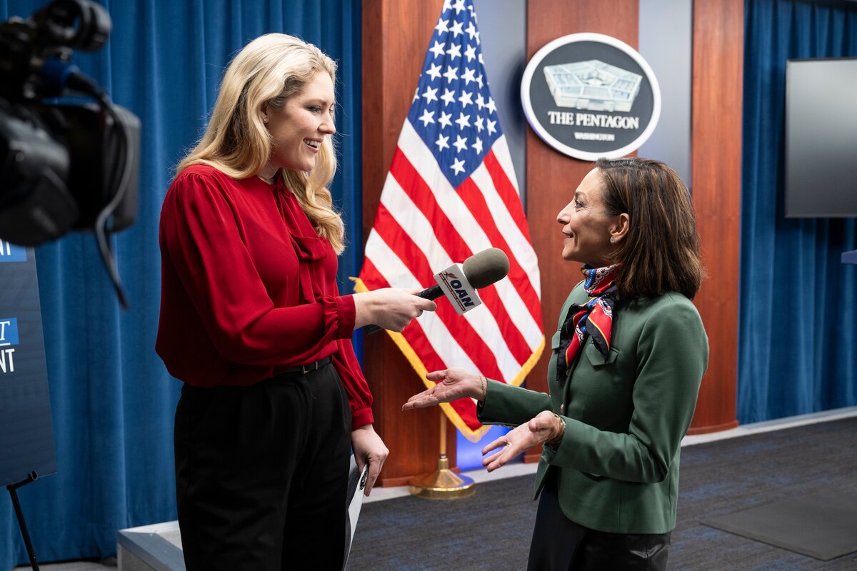 A person holding a microphone speaks with another person indoors, with an American flag and a sign that says "The Pentagon" in the background.