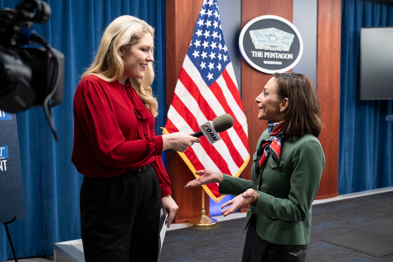 A person holding a microphone speaks with another person indoors, with an American flag and a sign that says "The Pentagon" in the background.