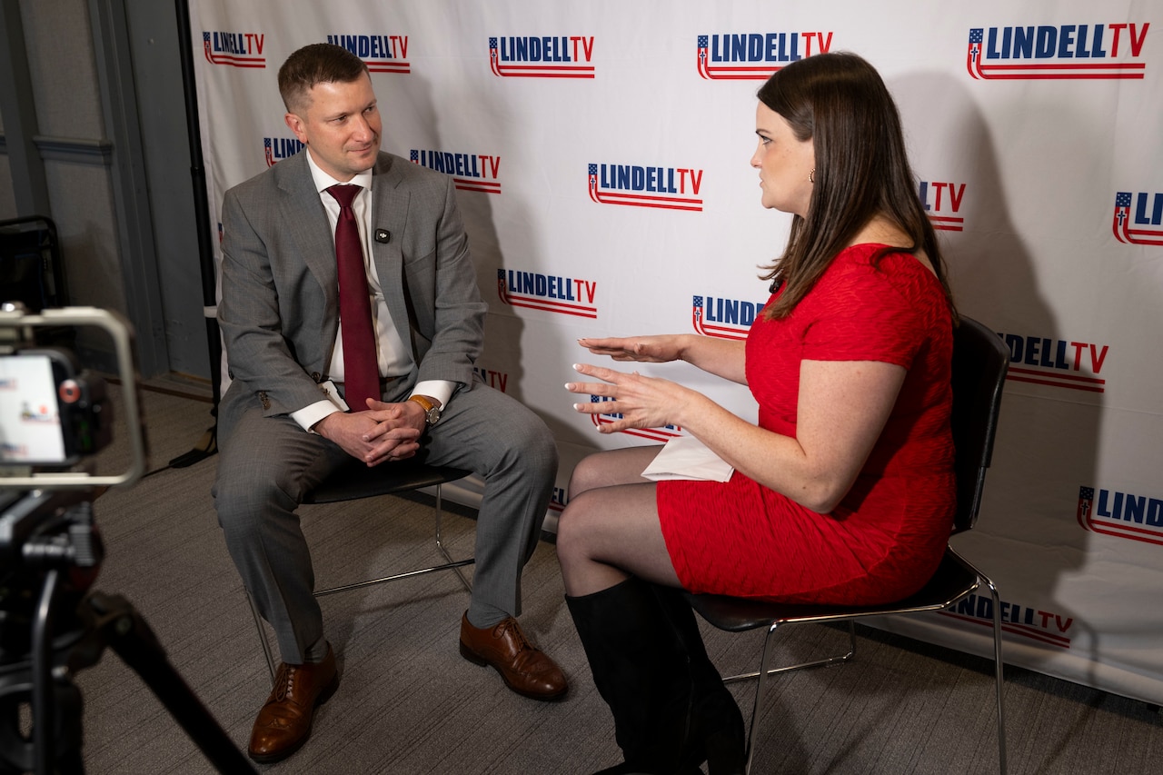 Two people sit on chairs facing one another in front of a screen that says "LindellTV."