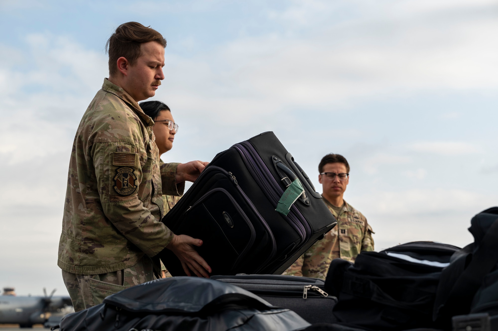 A military member puts a black suitcase on top of other bags.