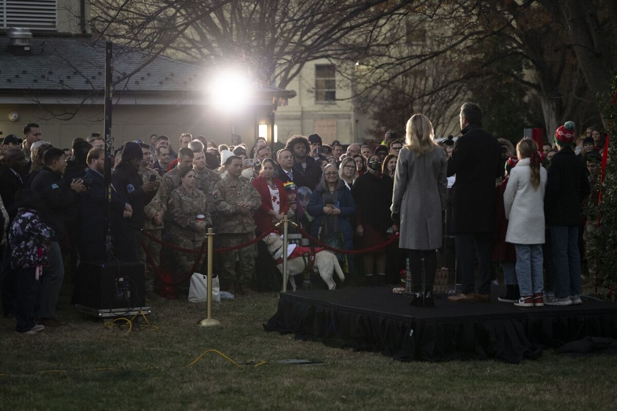 A large number of men and women (some in military uniform and some in civilian attire) as well as some children are standing outside at twilight, observing a man, a woman and children who are standing on a stage in front of them with their backs to the camera.