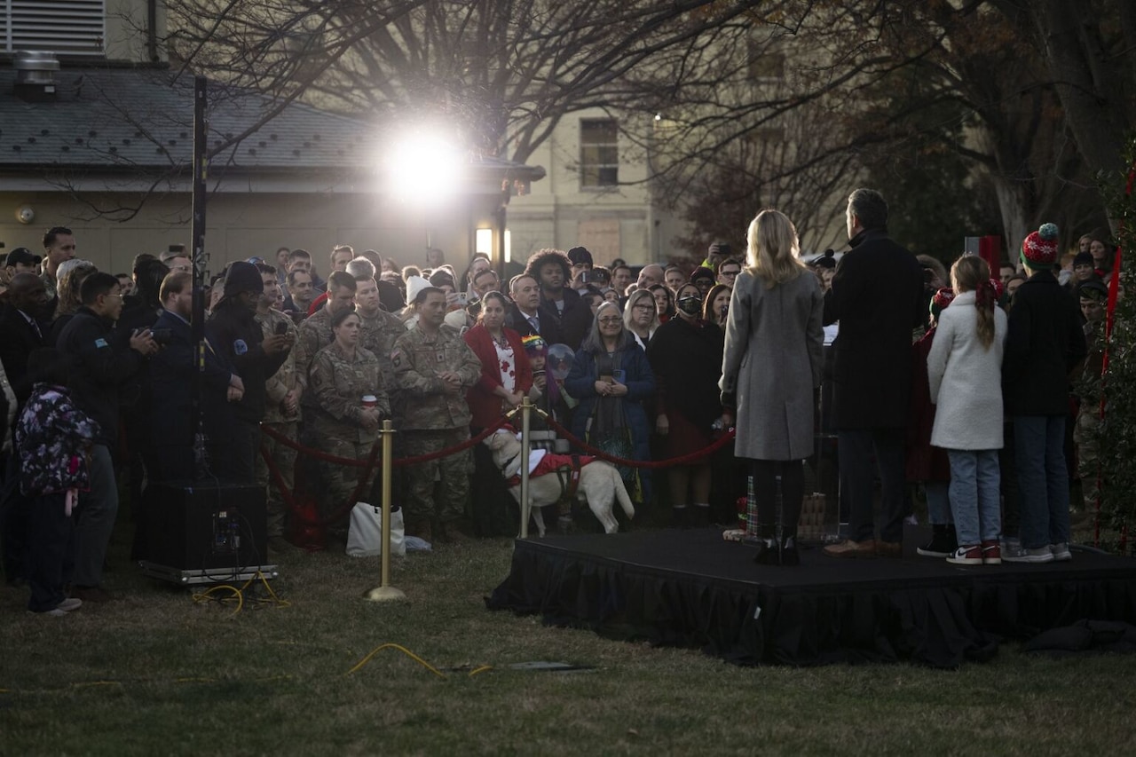 A large number of men and women (some in military uniform and some in civilian attire) as well as some children are standing outside at twilight, observing a man, a woman and children who are standing on a stage in front of them with their backs to the camera.
