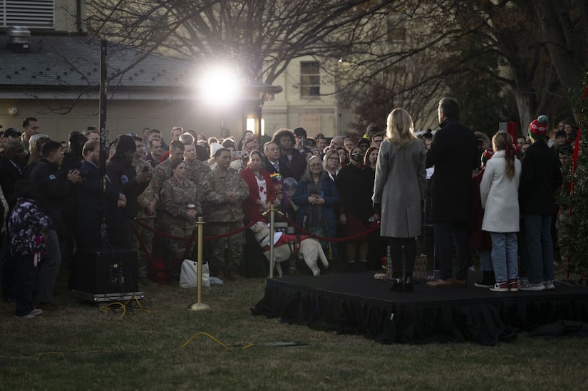 A large number of men and women (some in military uniform and some in civilian attire) as well as some children are standing outside at twilight, observing a man, a woman and children who are standing on a stage in front of them with their backs to the camera.