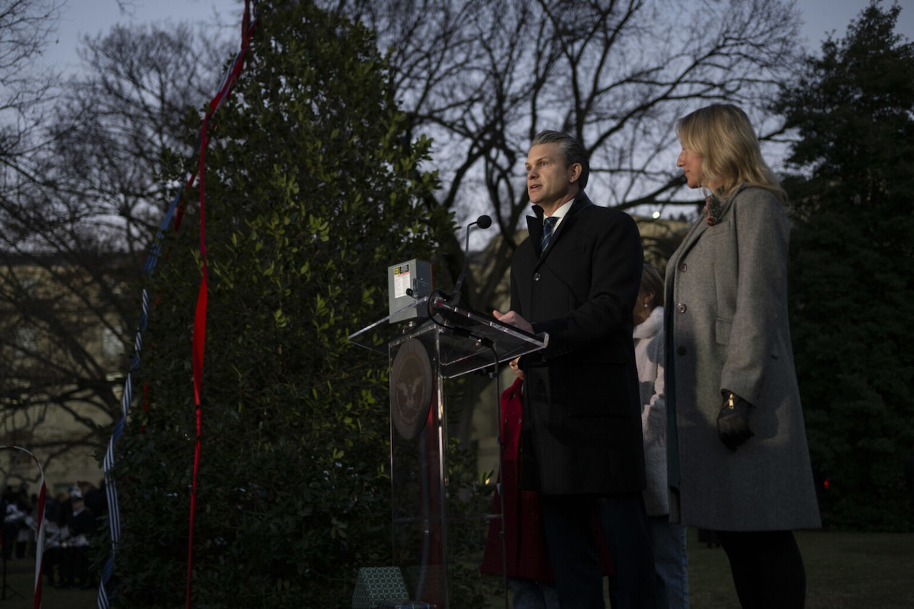 A man in a long winter coat is standing on a stage and speaking from behind a lectern into a microphone. There is a 14-foot-tall, unlighted Christmas tree to the left of him and woman in a winter coat to the right of him.