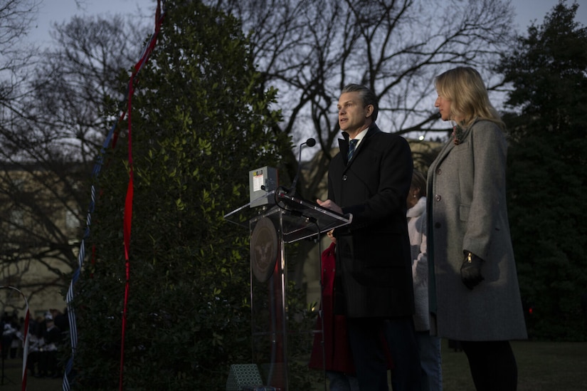 A man in a long winter coat is standing on a stage and speaking from behind a lectern into a microphone. There is a 14-foot-tall, unlighted Christmas tree to the left of him and woman in a winter coat to the right of him.