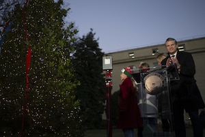 A person in a long winter coat is standing on a stage behind a podium at twilight, smiling and clapping. A child stands nearby, looking at a 14-foot-tall, lighted Christmas tree
