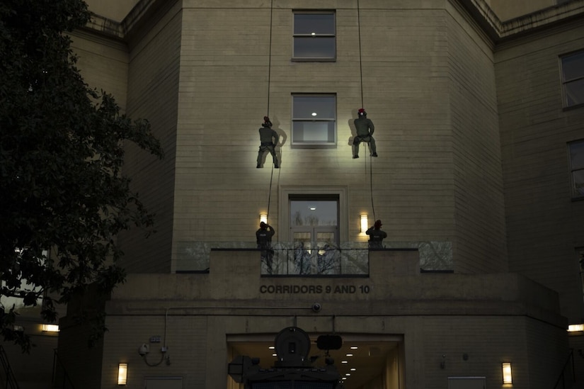Four people with their backs to the camera are rappelling down the side of a five-story building at twilight. There are two people to each line. A sign on the first floor of the building reads "corridors 9 and 10."