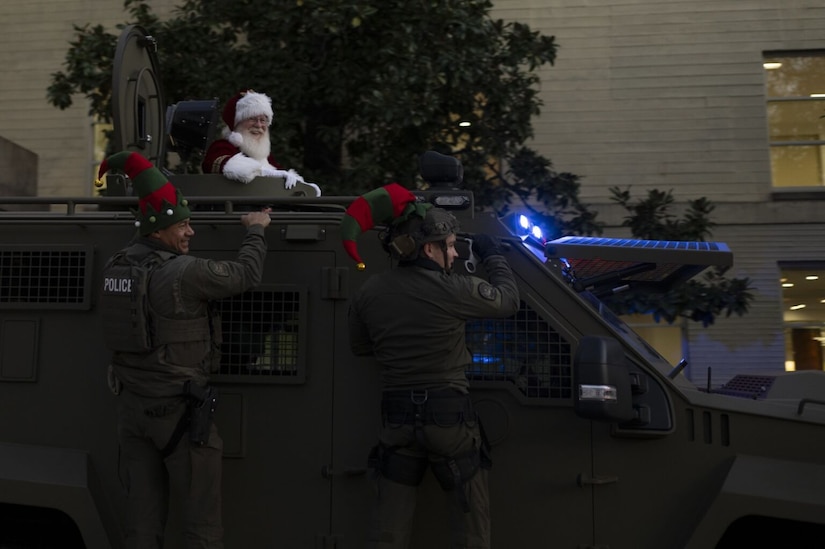 A man dressed as Santa Claus rides in the center of an armored tactical vehicle at twilight. Two men in tactical police gear and elf hats are mounted standing on the vehicles side with their backs to the camera.