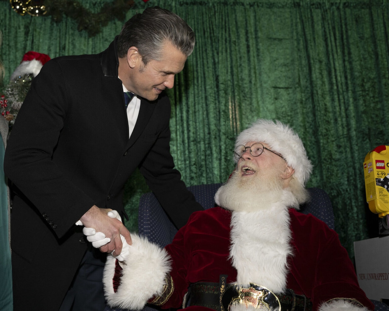 A person in civilian winter business attire is leaning over and shaking the hand of a seated person dressed as Santa Claus. They are both smiling.