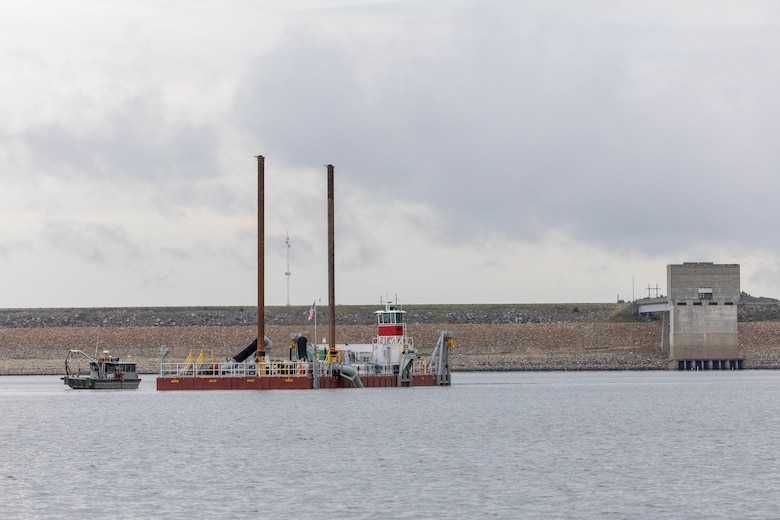 A large barge with two brown smokestacks sits in water with a dam outlet tower to the right, rocks and grey sky in the background.