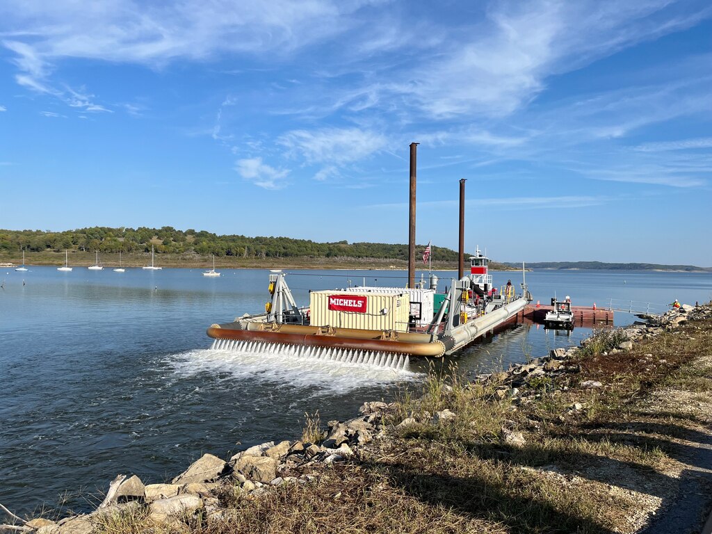 A large barge with two brown smokestacks sits in water with rock in the foreground, vegetation and blue sky in the background.