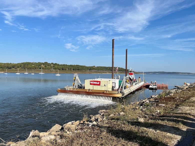 A large barge with two brown smokestacks sits in water with rock in the foreground, vegetation and blue sky in the background.