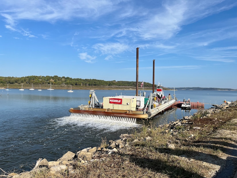 A large barge with two brown smokestacks sits in water with rock in the foreground, vegetation and blue sky in the background.