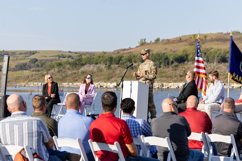 A man in an Army uniform speaks at a podium in front of a large audience with an American flag to the right, water and vegetation in the background.