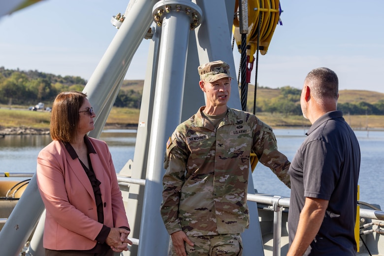 A woman in a pink blazer, a man in an Army uniform, and a man in a grey shirt talk with water and vegetation in the background.