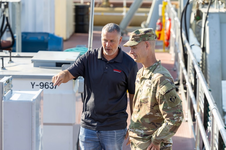 A man in a grey polo and a man in an Army uniform look at something out of frame to the left.
