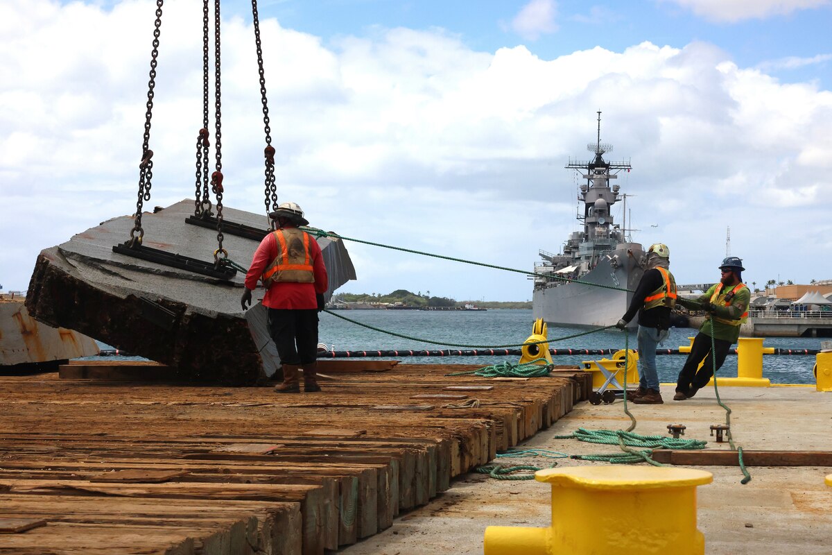 Two people wearing reflective vests and hard hats pull on ropes attached to a large concrete slab hanging from chains. Another person in similar attire stands next to the concrete slab. There is a large military ship docked in the background.