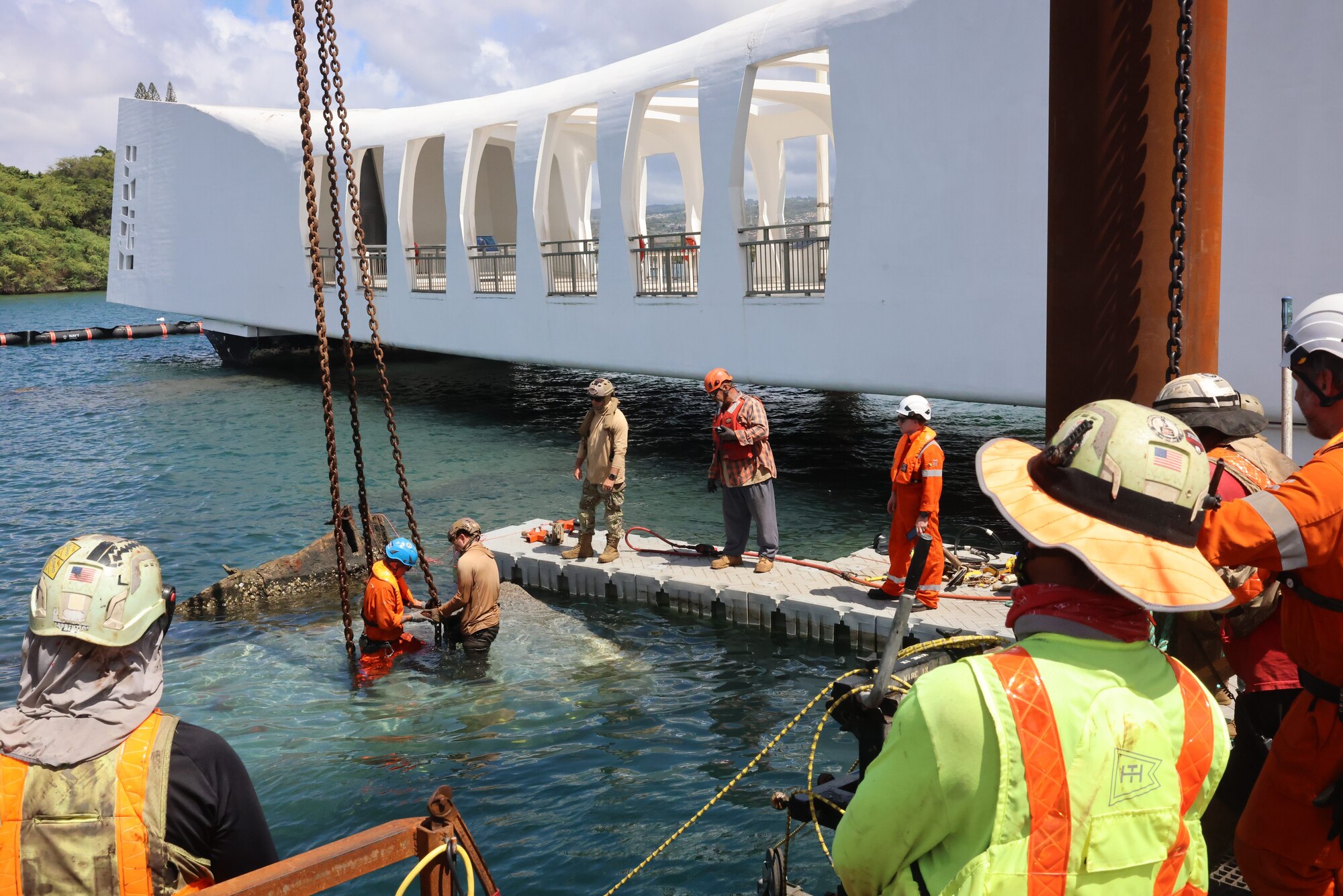 Two people wearing hard hats attach chains to a large concrete slab in the water. Several other people in similar attire stand on platforms, observing the two people in the water. There is a large floating, white structure in the background.