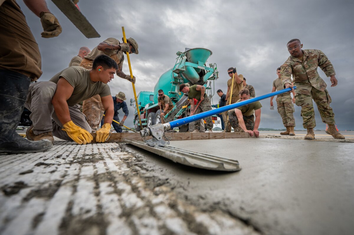 Airmen dressed in military camouflage uniforms spread concrete on a runway.
