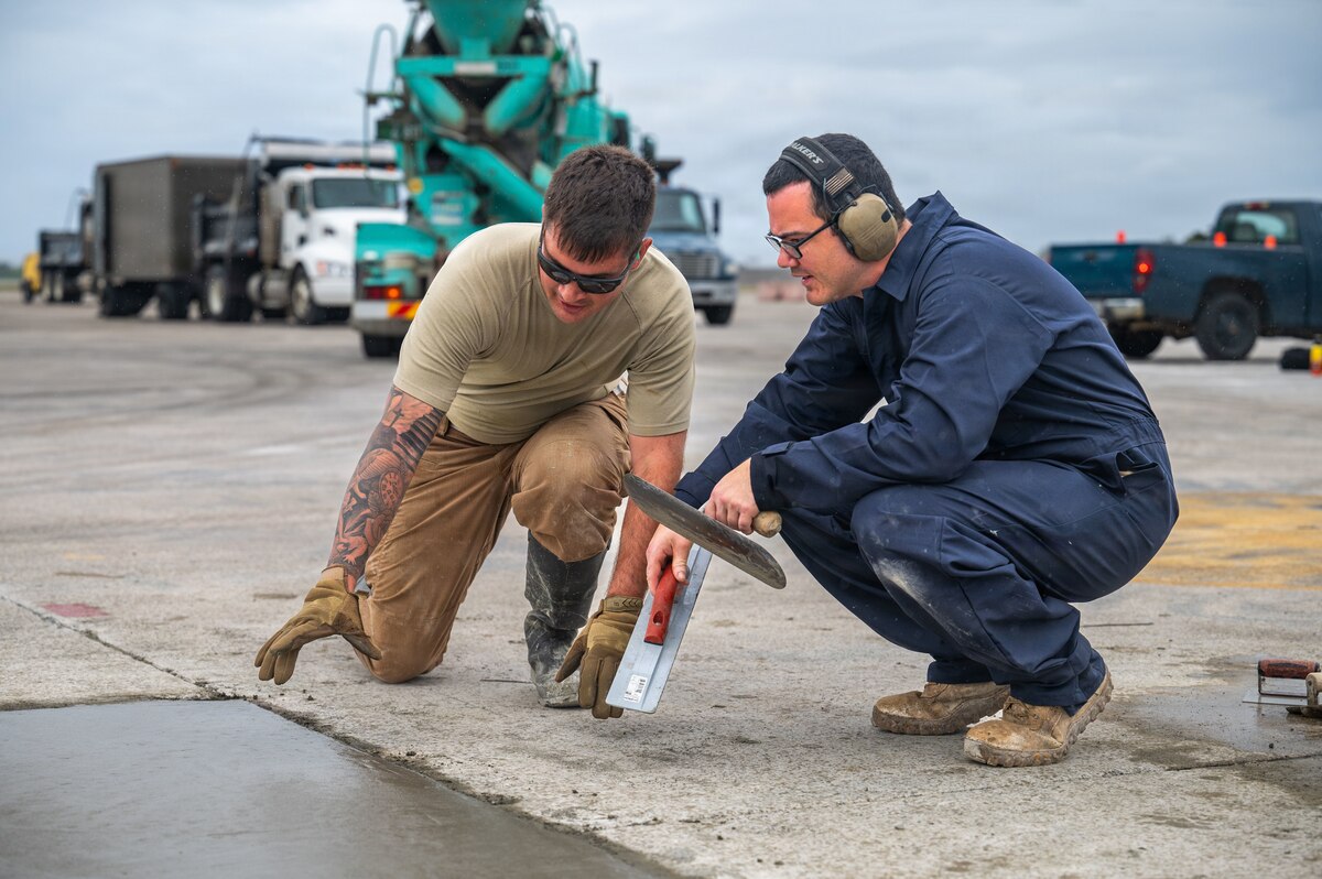 A man on the left wearing sunglasses, a tan T-shirt and pants, and rubber boots kneels next to a freshly poured patch of concrete while speaking to a man to his right who is also kneeling and wearing blue coveralls, hearing protection and glasses.