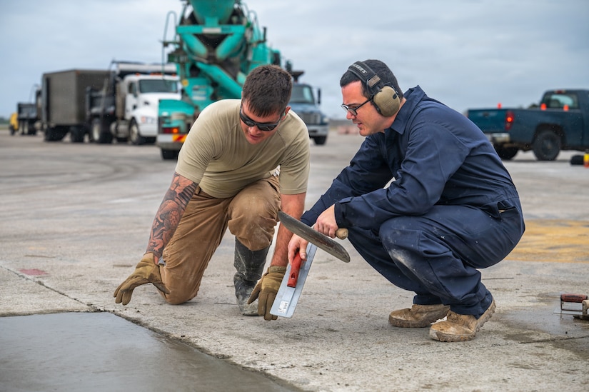A man on the left wearing sunglasses, a tan T-shirt and pants, and rubber boots kneels next to a freshly poured patch of concrete while speaking to a man to his right who is also kneeling and wearing blue coveralls, hearing protection and glasses.