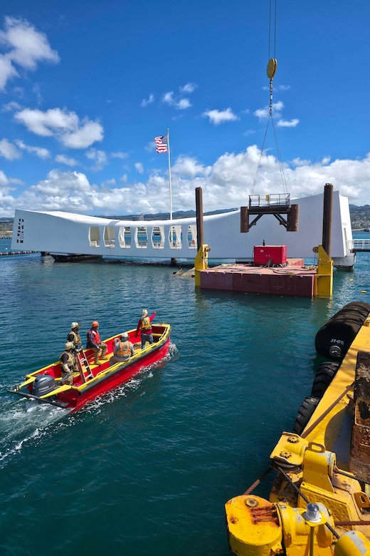 Five people ride in a boat toward a platform in the water. A large crane is lifting a metal object from the platform. There is a large floating white structure in the background with an American flag flying from the top.