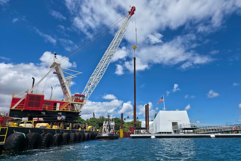 A crane sitting on a barge moves metal pillars into the water. There is a large floating white structure in the background, with an American flag flying from the top.