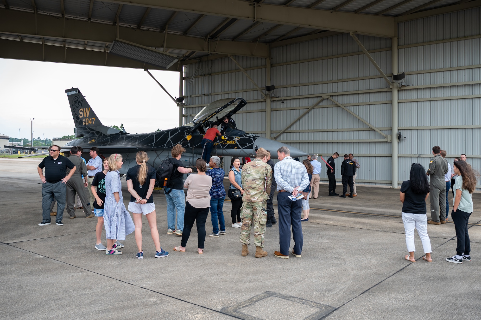 Civilians stand on a runway apron and look at a fighter jet under a hangar.