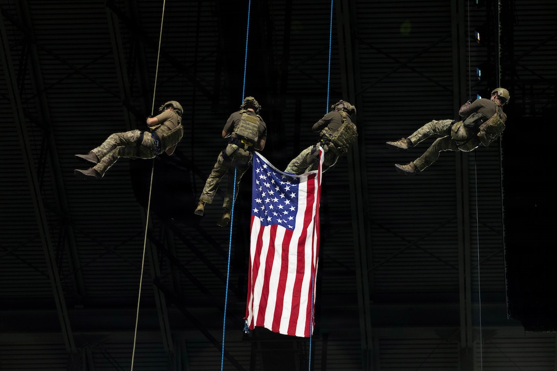Four soldiers, two with the corners of an American flag attached to their gear, rappel from a large structure.