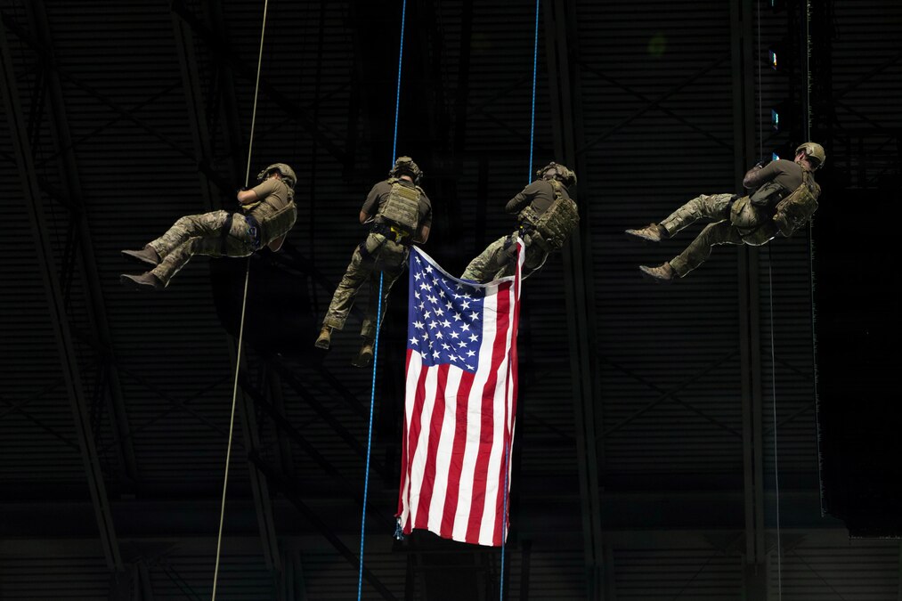 Four soldiers, two with the corners of an American flag attached to their gear, rappel from a large structure.