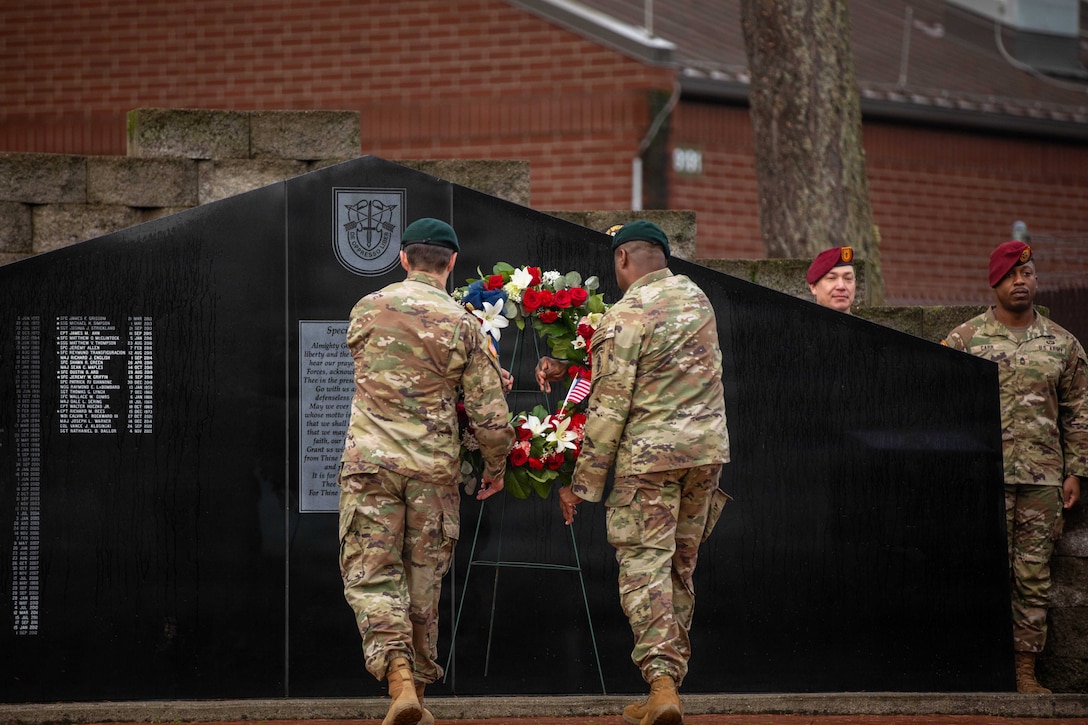 Two soldiers hold a wreath display and walk toward a black memorial outdoors, with two service members standing behind the memorial and a brick building in the background.