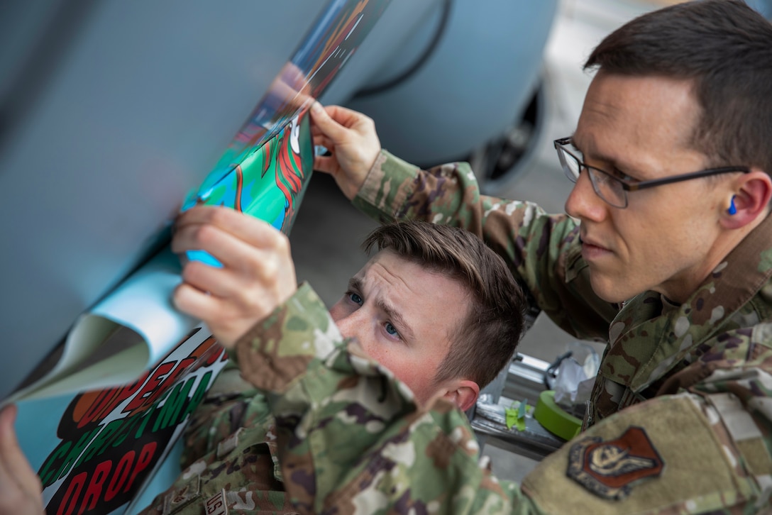 Two airmen wearing camouflage apply a large decal to an aircraft.