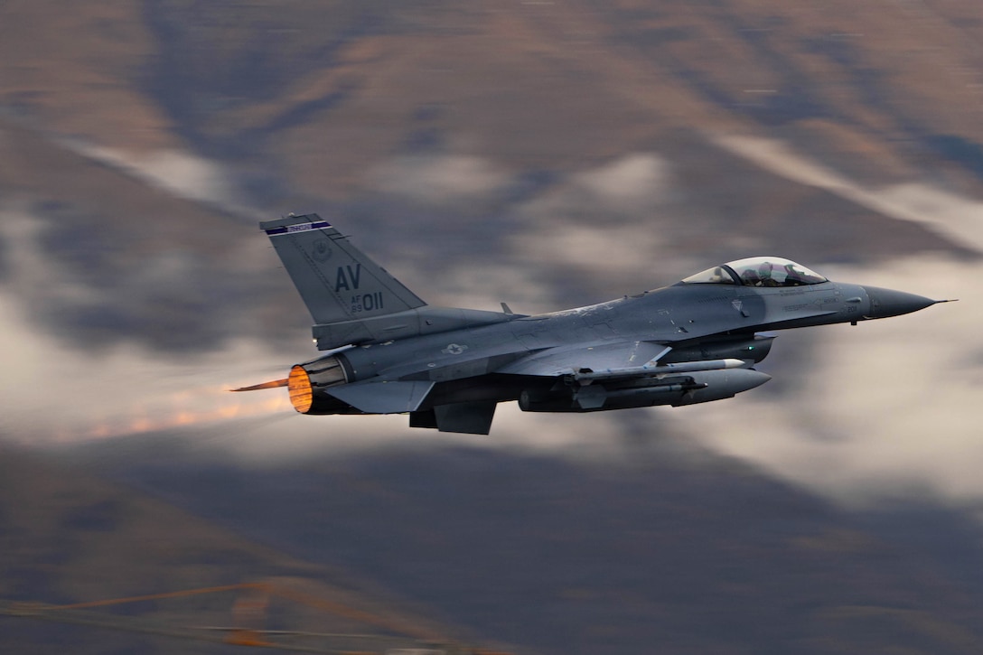 A fighter jet accelerates in the sky during daytime, with mountainous land and clouds in the background.
